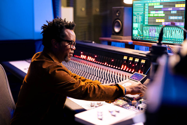 Afrikai amerikai zenei producer zeneszerzés számokat a vezérlőteremben, vágás dalok és hangerő beállítások műszerfal gombokkal. Hangmérnök nyomkodja a fadereket és gombokat. - Fotó, kép