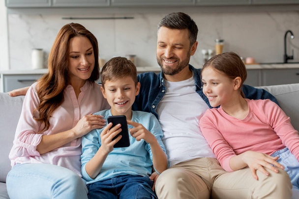 Μια χαρούμενη οικογένεια με τέσσερις αγκαλίτσες στον καναπέ και κοιτάζοντας ένα smartphone, επιδεικνύοντας μια στιγμή δεσίματος και τεχνολογικής αλληλεπίδρασης - Φωτογραφία, εικόνα