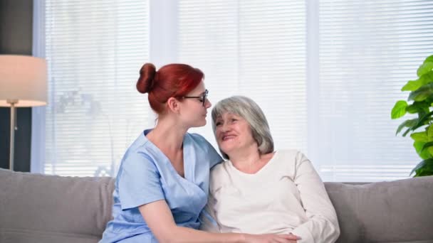 asistencia social para personas mayores, una niñera joven con uniforme médico apoya a una anciana sentada en el sofá en la habitación - Imágenes, Vídeo