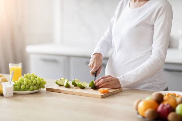 Těhotná evropská dáma v bílé košili připravuje ve své moderní kuchyni výživný ovocný salát, který ztělesňuje zdraví a sebepéči - Fotografie, Obrázek