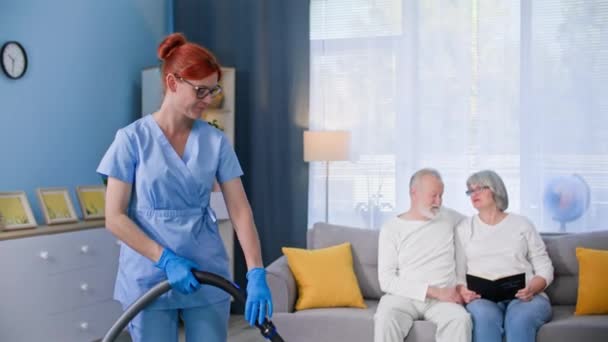 helping pensioners, young female nurse in a medical suit helps with cleaning old woman and an elderly man vacuuming floor in room - Footage, Video