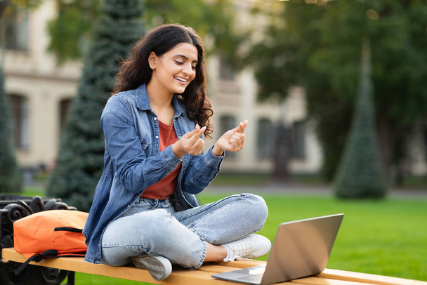 Positive junge indische Studentin hat Videoanruf mit Klassenkameradin im Freien, Blick auf Laptop-Bildschirm, zeigt Liebesgeste - Foto, Bild