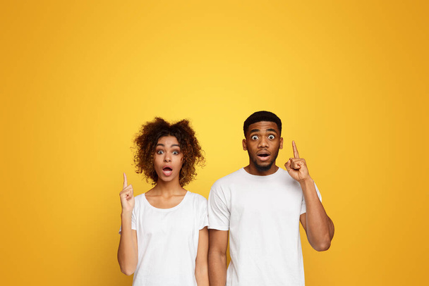 Verbijsterd zwarte man en vrouw naar boven wijzen met geopende mond in verbazing, oranje achtergrond - Foto, afbeelding