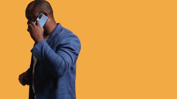 Αφροαμερικάνος που έχει σημαντικό τηλεφώνημα με τον συνεργάτη του χρησιμοποιώντας smartphone, φόντο στούντιο. BIPOC πρόσωπο με τα πόδια γύρω συζητώντας θέματα της εταιρείας κατά τη διάρκεια τηλεφωνικής κλήσης, κάμερα Β - Πλάνα, βίντεο
