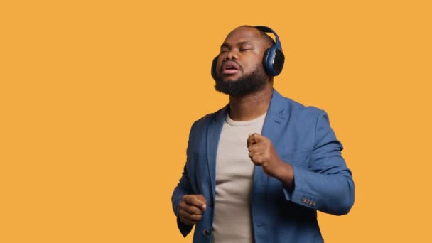 Boldog afro-amerikai ember szórakozik, ritmusra táncol, fejhallgatót hallgat. Vidám BIPOC férfi szórakoztatja magát csinál funky mozgások zene, stúdió háttér, kamera A - Felvétel, videó