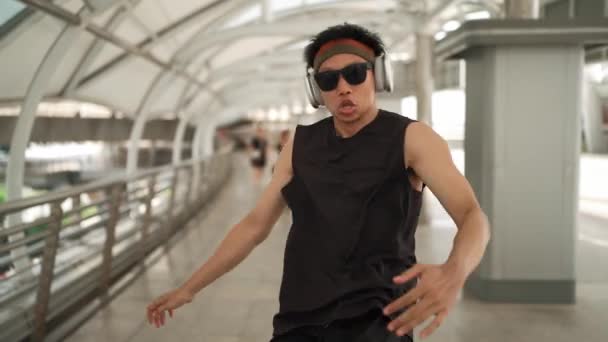 Mądry przystojny tancerz porusza rękami podczas tańca do hip-hopu. Profesjonalny azjatycki hipster noszący słuchawki podczas spaceru korytarzem z zamazanym tłem. Sport na świeżym powietrzu 2024. Sprytnie. - Materiał filmowy, wideo