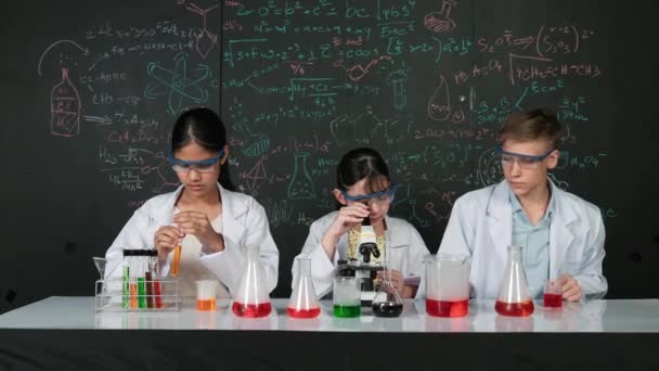 Schattig meisje kijkt onder de microscoop terwijl de student experimenteert op schoolbord met theorie geschreven. Jonge wetenschapper inspecteren gekleurde oplossing aan tafel met experimentele apparatuur geplaatst. Edificatie - Video