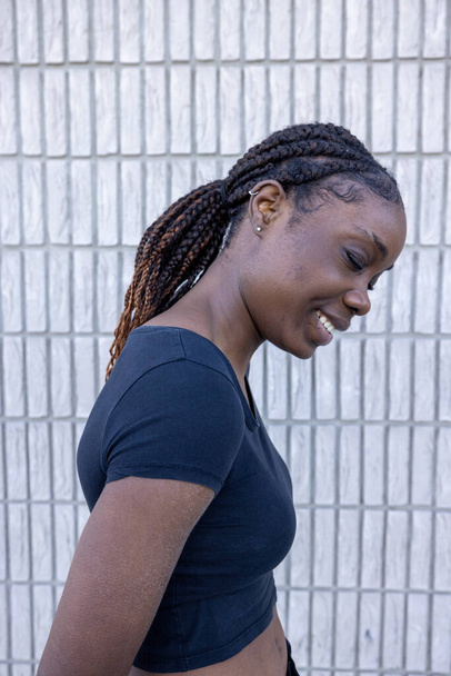 Capturada de perfil, esta imagen representa a una joven africana con una sonrisa alegre, mirando hacia abajo. Su cabello está peinado con elegantes trenzas que cubren su hombro, y lleva un negro casual. - Foto, Imagen