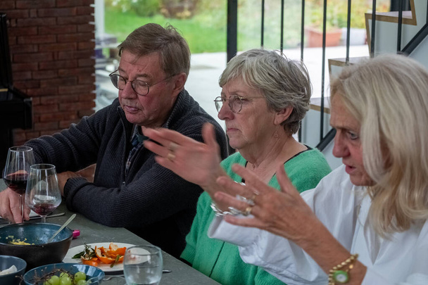 Un'istantanea intima di un gruppo di amici anziani profondamente coinvolti in una conversazione durante un pasto. L'ambiente è accogliente e informale, con piatti e bicchieri che indicano un'esperienza di cena condivisa. A - Foto, immagini