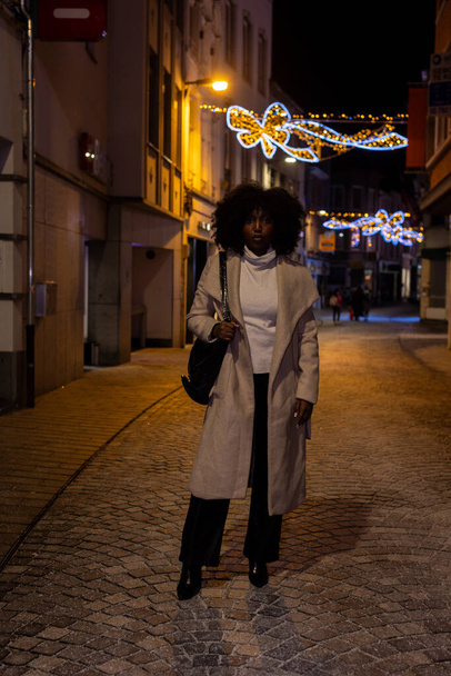 Nainen ottaa kaupungin kaduilla raikas talvi-iltana, hänen polku valaistu juhlava valot koukussa edellä. Hänen muoti on sekoitus vuodenajan lämpöä ja modernia tyyliä, pitkä takki kerroksellinen - Valokuva, kuva