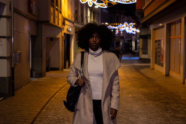 Esta imagem impressionante captura uma mulher confiante caminhando em direção à câmera em uma rua de paralelepípedos vazia à noite. Sua negrito afro e roupa elegante são iluminados pelo brilho suave de lâmpadas de rua - Foto, Imagem