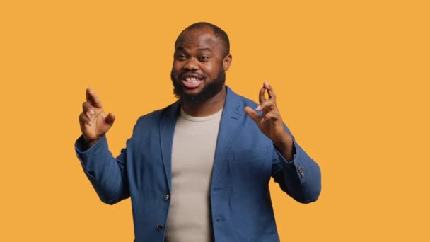 Portret van een vrolijke Afrikaans-Amerikaanse man die zijn vingers kruist, wensend, geïsoleerd over een gele studio achtergrond. Jolly BIPOC persoon in de hoop op veel geluk, wachten op wonder, camera A - Video