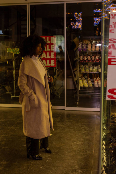 Egy városi este melege van beburkolva ebbe a képbe, ahol egy nő áll egy kirakatnál, az ELADÓ szó félkövér betűkkel tükröződik. Az ünnepi fények csillognak a háttérben, ami arra utal, egy - Fotó, kép