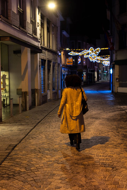 この画像は,ストリートライトの柔らかい光の下の石畳の通りに沿って歩いている孤独な姿を示し,遠くに輝くお祝いのライト. 長いコートと自信を持っている人 - 写真・画像