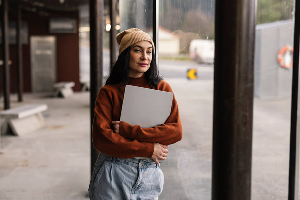 Молодая женщина с ноутбуком уверенно стоит на автобусной станции, воплощая городскую мобильность и внештатный образ жизни. - Фото, изображение