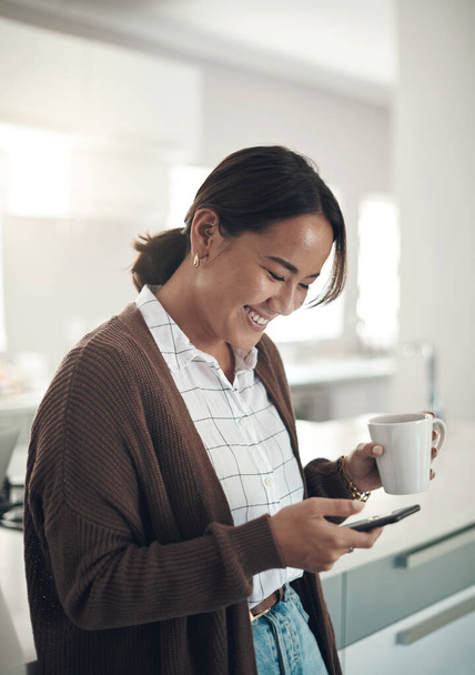 Teléfono, sonrisa y mujer asiática tomando café en la cocina en casa para el desayuno por la mañana. Smartphone, taza de té y persona feliz en las redes sociales, búsqueda en Internet y correo electrónico de lectura en la aplicación móvil. - Foto, imagen