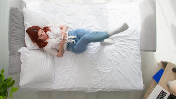 relaxant à la maison, jeune femme utilise smartphone pour naviguer sur Internet, commande des marchandises en ligne tout en étant couché sur le lit, vue de dessus - Séquence, vidéo