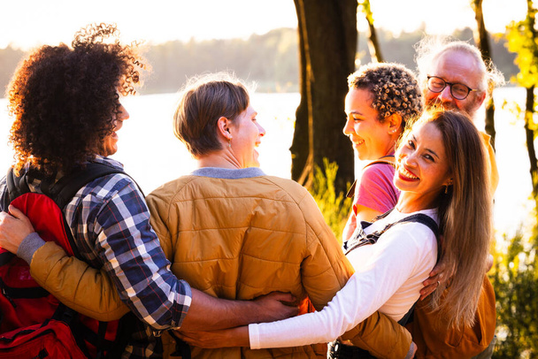 Αυτή η συγκινητική εικόνα αιχμαλωτίζει μια οικογένεια πολλών γενεών απολαμβάνοντας μια στιγμή συντροφικότητας δίπλα σε μια λίμνη το ηλιοβασίλεμα. Η ομάδα συμμετέχει σε μια ομαδική αγκαλιά, με χαρούμενες εκφράσεις και μια αύρα - Φωτογραφία, εικόνα