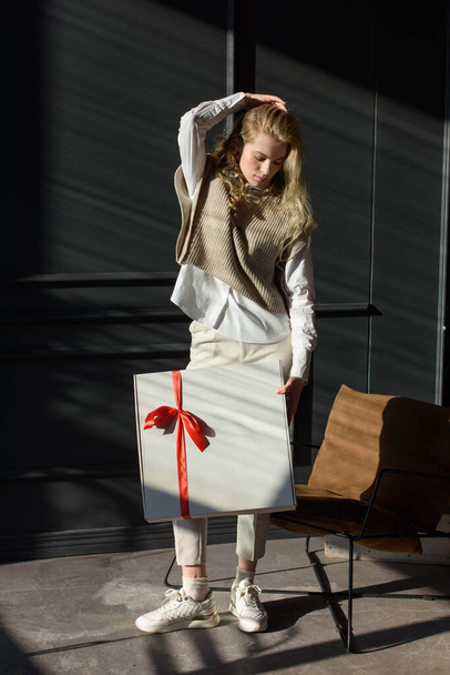Naisella on valkoinen lahjapakkaus, jossa on punainen nauha. Malli yllään tyylikäs neulottu liivi, valkoinen paita ja klassiset housut. - Valokuva, kuva
