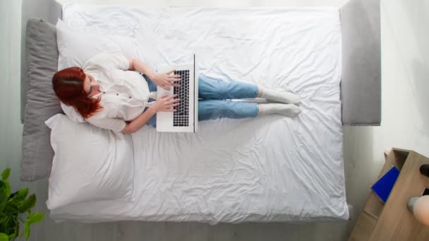 vrouw maakt gebruik van laptop om te communiceren met vrienden of werk terwijl liggend op bed in de kamer, bovenaanzicht - Video