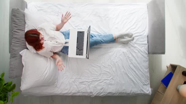 młoda pogodna kobieta odpoczywająca na łóżku z komputerem w ręku komunikuje się z przyjaciółmi przez Internet za pomocą kamery internetowej, widok z góry - Materiał filmowy, wideo
