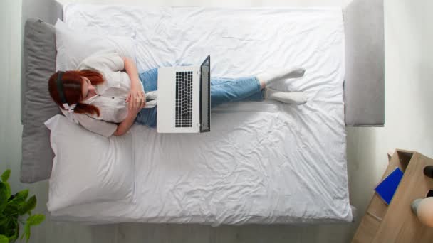 mujer joven con auriculares hablando a través de videoconferencia en el ordenador utilizando la cámara web mientras se relaja en la cama en el dormitorio, vista superior - Metraje, vídeo
