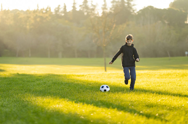 少年は野原でサッカーをしている. 草は緑色で,空は青色になっています. 少年は黒いジャケットとブルージーンズを着ています - 写真・画像