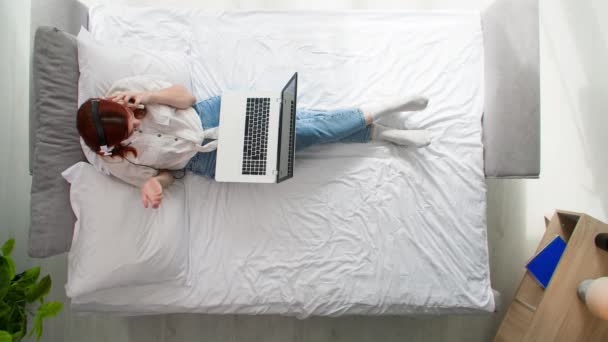 работая дома, женщина использует портативную гарнитуру для общения с друзьями или работы, лежа на кровати в комнате, вид сверху - Кадры, видео