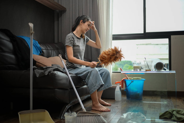 Jonge huisvrouw zit op de bank met schoonmaakmiddelen gevoel moe na het doen van huishoudelijk werk. - Foto, afbeelding