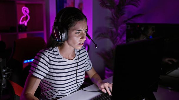 Νεαρή όμορφη ισπανόφωνη γυναίκα streamer φορώντας ακουστικά παίζοντας βιντεοπαιχνίδι στην αίθουσα τυχερών παιχνιδιών - Φωτογραφία, εικόνα