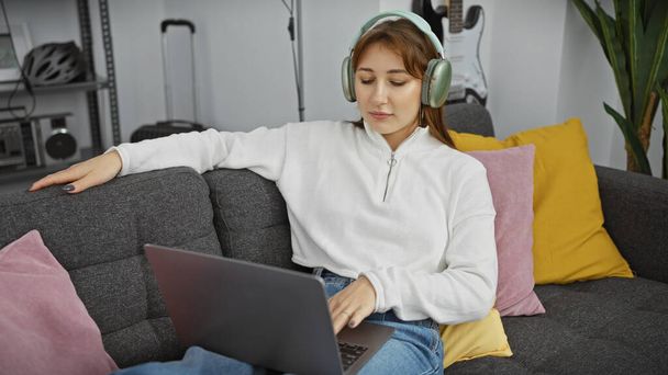 Zrelaksowana młoda kobieta nosząca słuchawki podczas używania laptopa na kanapie w pomieszczeniu. - Zdjęcie, obraz