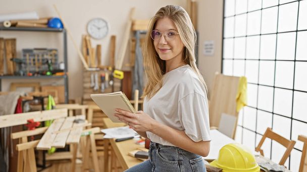 Portrait of a young adult caucasian woman with blonde hair, wearing eyeglasses, casually dressed, standing in a workshop holding a notebook. - Photo, Image