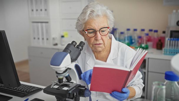 Femme scientifique chevronnée lisant un livre tout en travaillant au microscope en laboratoire. - Photo, image