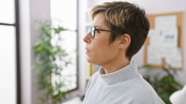 A contemplative young hispanic woman with short blonde hair wearing glasses and a turtleneck indoors an office setting. - Photo, Image