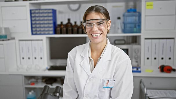Αυτοπεποίθηση και ευτυχία, νέα, όμορφη Ισπανίδα επιστήμονας, χαμογελώντας, απολαμβάνοντας τη δουλειά της στο εργαστήριο με γυαλιά ασφαλείας - ένα πορτρέτο εμπιστοσύνης και χαράς στην επιστήμη - Φωτογραφία, εικόνα