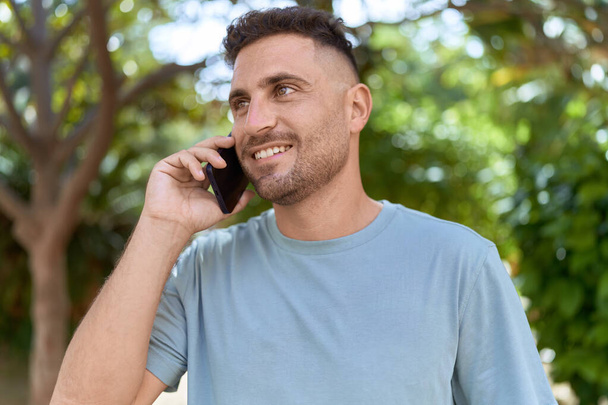 Genç İspanyol adam gülümsüyor. Parktaki akıllı telefondan konuşuyor. - Fotoğraf, Görsel
