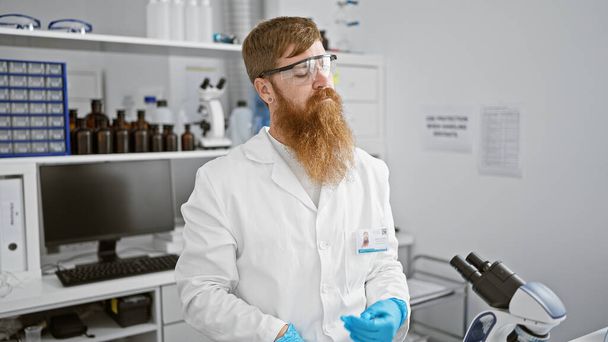 Расслабленный, но серьезный молодой рыжий ученый мужского пола, погруженный в новаторский эксперимент в лаборатории, обеспечивает безопасность в защитных очках - Фото, изображение