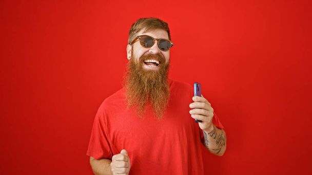 Веселий молодий ірландський чоловік з бородою, танцює над ізольованою червоною стіною, радісно слухаючи смішні мелодії на мобільному телефоні. красивий рудий хлопець посміхається, тримає мобільний телефон, насолоджуючись онлайн музикою. - Фото, зображення
