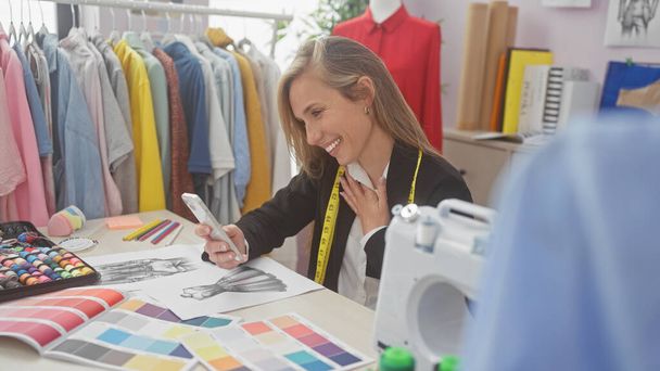 Uśmiechnięta kobieta korzystająca ze smartfona w kolorowym studiu mody z tkaniną i maszyną do szycia - Zdjęcie, obraz