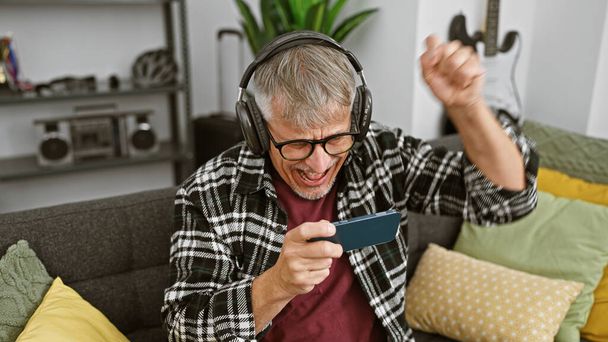 Izgalmas idős férfi fejhallgató játszik okostelefon játék kanapén otthon - Fotó, kép