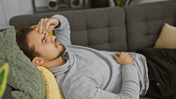 Ισπανόφωνος άντρας χαλαρώνει στον καναπέ στο σπίτι με το χέρι στο μέτωπο, δείχνοντας αγχωμένος ή κουρασμένος. - Φωτογραφία, εικόνα