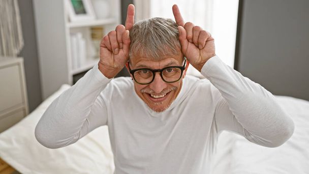 Homme d'âge moyen souriant aux cheveux gris faisant un geste d'oreilles de lapin assis sur un lit blanc dans une chambre moderne. - Photo, image