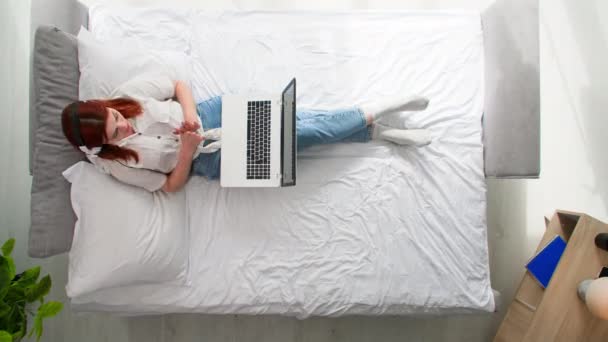 жінка з гарнітурою і смартфоном працює на ноутбуці, лежачи на ліжку в спальні, вид зверху - Кадри, відео
