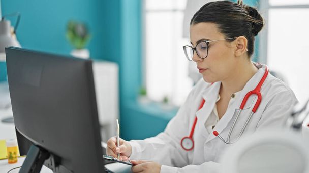 Νεαρή όμορφη ισπανόφωνη γιατρός χρησιμοποιώντας το γράψιμο του υπολογιστή στο πρόχειρο στην κλινική - Φωτογραφία, εικόνα