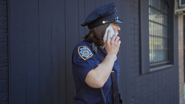 Ώριμη γυναίκα αστυνομικός με στολή μιλάει στο τηλέφωνο έξω στο αστικό περιβάλλον. - Φωτογραφία, εικόνα