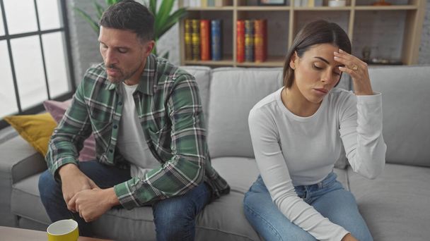 Een bezorgd Spaans echtpaar zit apart op de bank in een moderne woonkamer, wat de spanning en bezorgdheid weerspiegelt.. - Foto, afbeelding