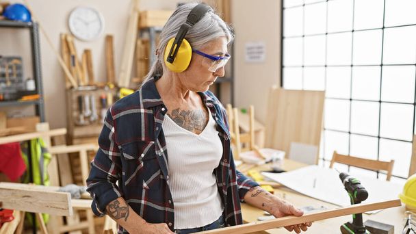 Serious, grey-haired middle age woman carpenter deeply invested in her woodwork, fervently looking at a timber plank in her homely carpentry workshop - Photo, Image
