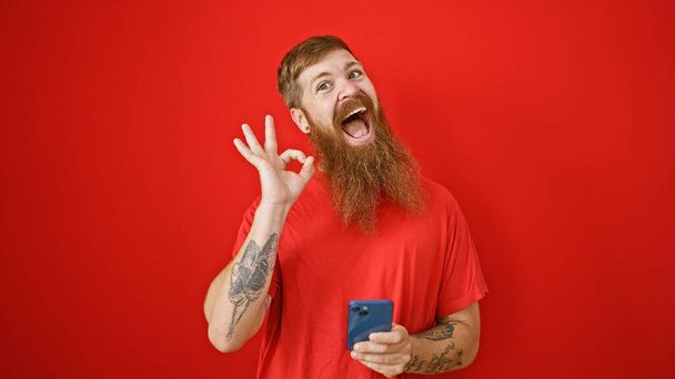 Stralende roodharige man geeft ok teken, terwijl verdiept in berichten op zijn smartphone, staat vol vertrouwen tegen een levendige rode muur, uitstralen vreugde, en positiviteit. - Foto, afbeelding