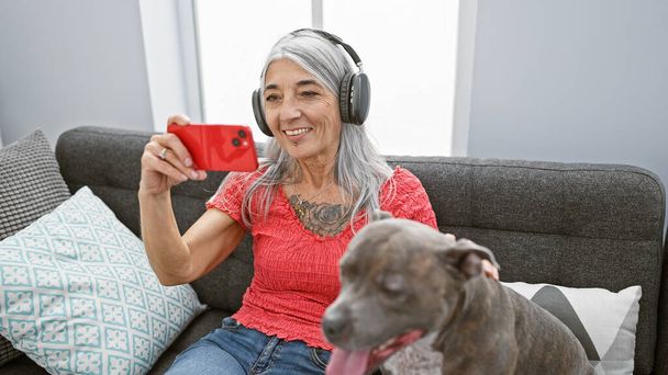 À la maison, une femme d'âge moyen aux cheveux gris se relaxant confortablement avec son chien, tout en utilisant son smartphone et son casque, sur son canapé - Photo, image