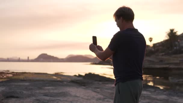 Egy férfi panorámás képet készít a tengerről naplementekor egy mobiltelefonon. Spanyolország, Cabo de Huertas. - Felvétel, videó